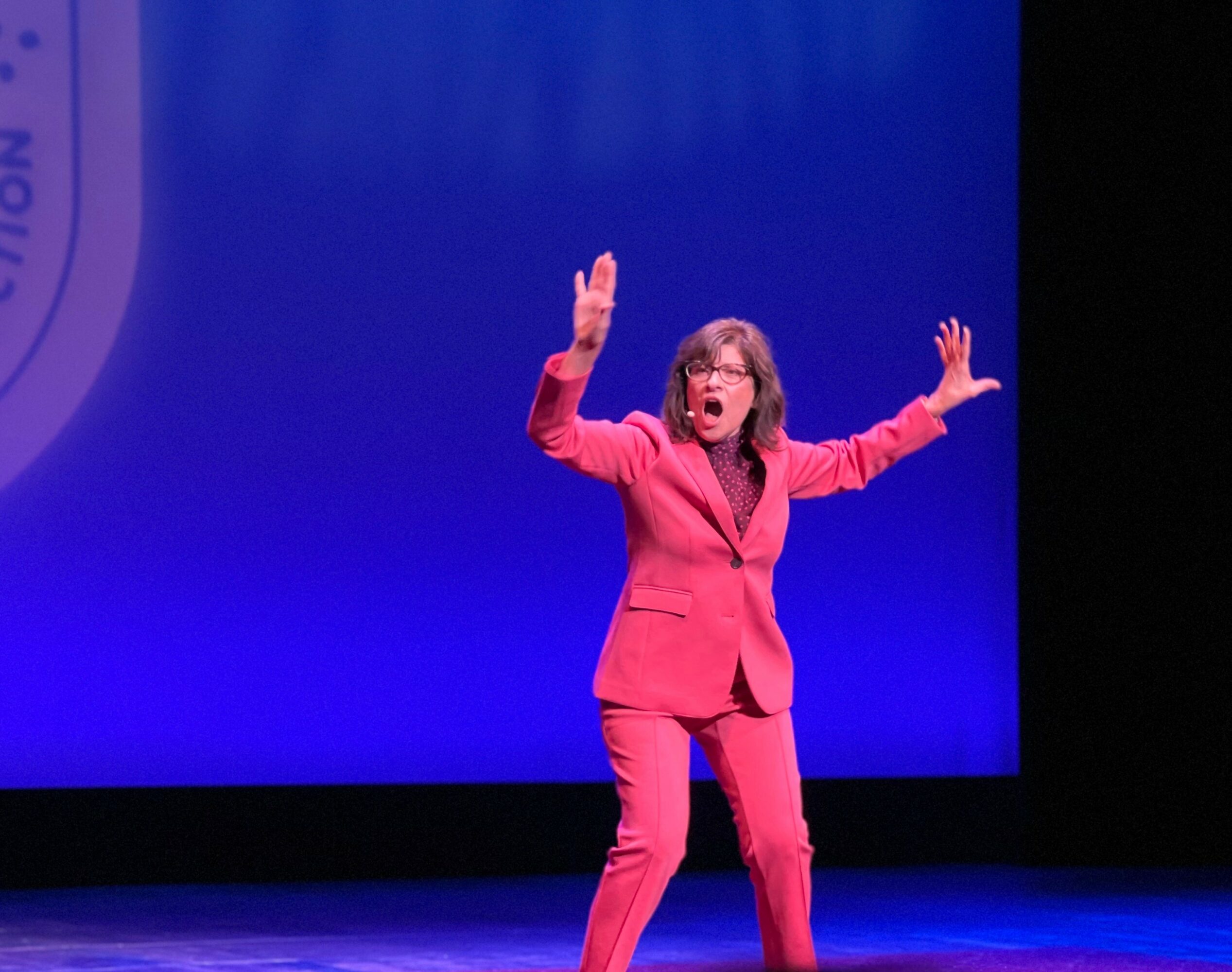 Woman in pink suit speaking on stage with blue background, gesturing with hands raised.