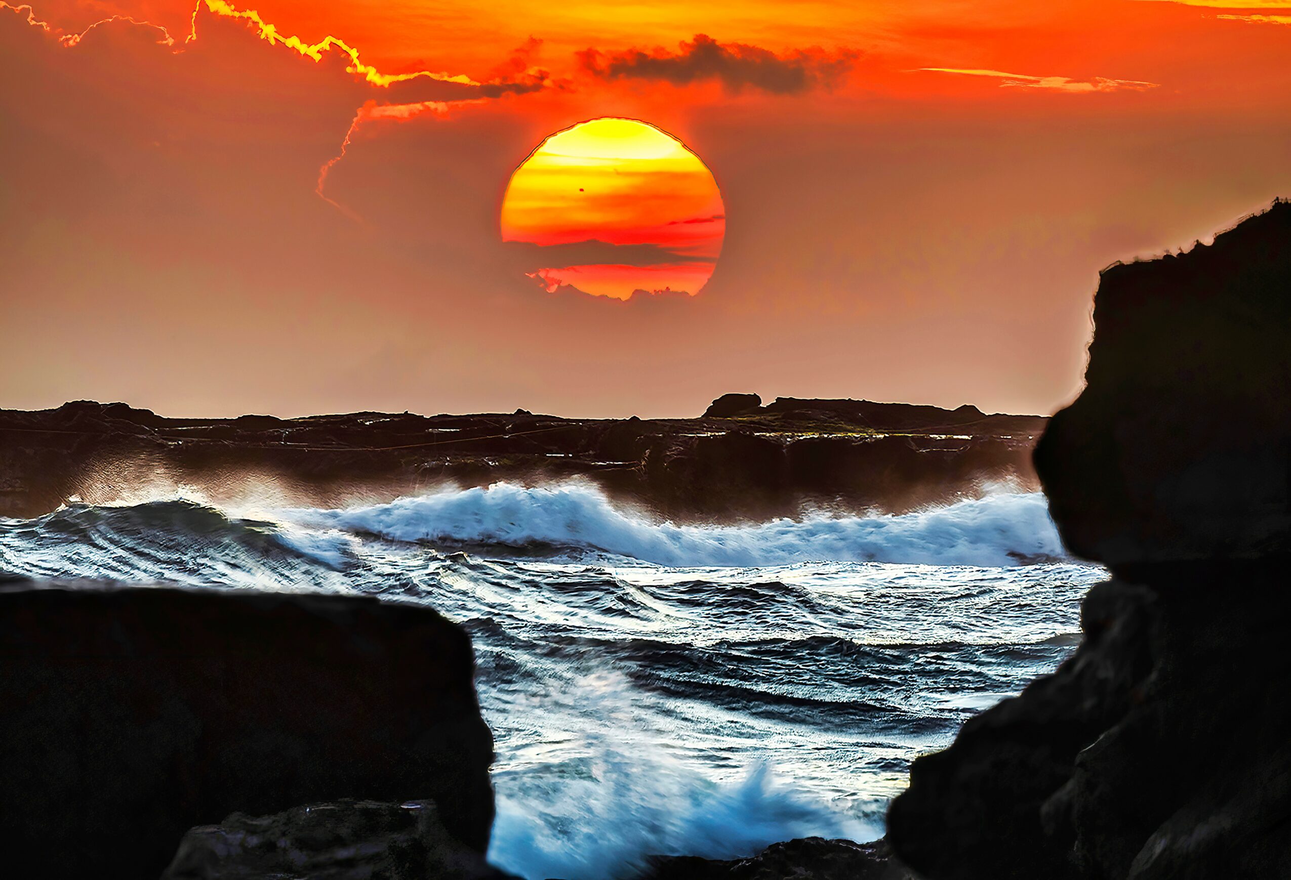 Sunset over ocean with waves crashing against rocks and a silhouette of a cliff on the right.