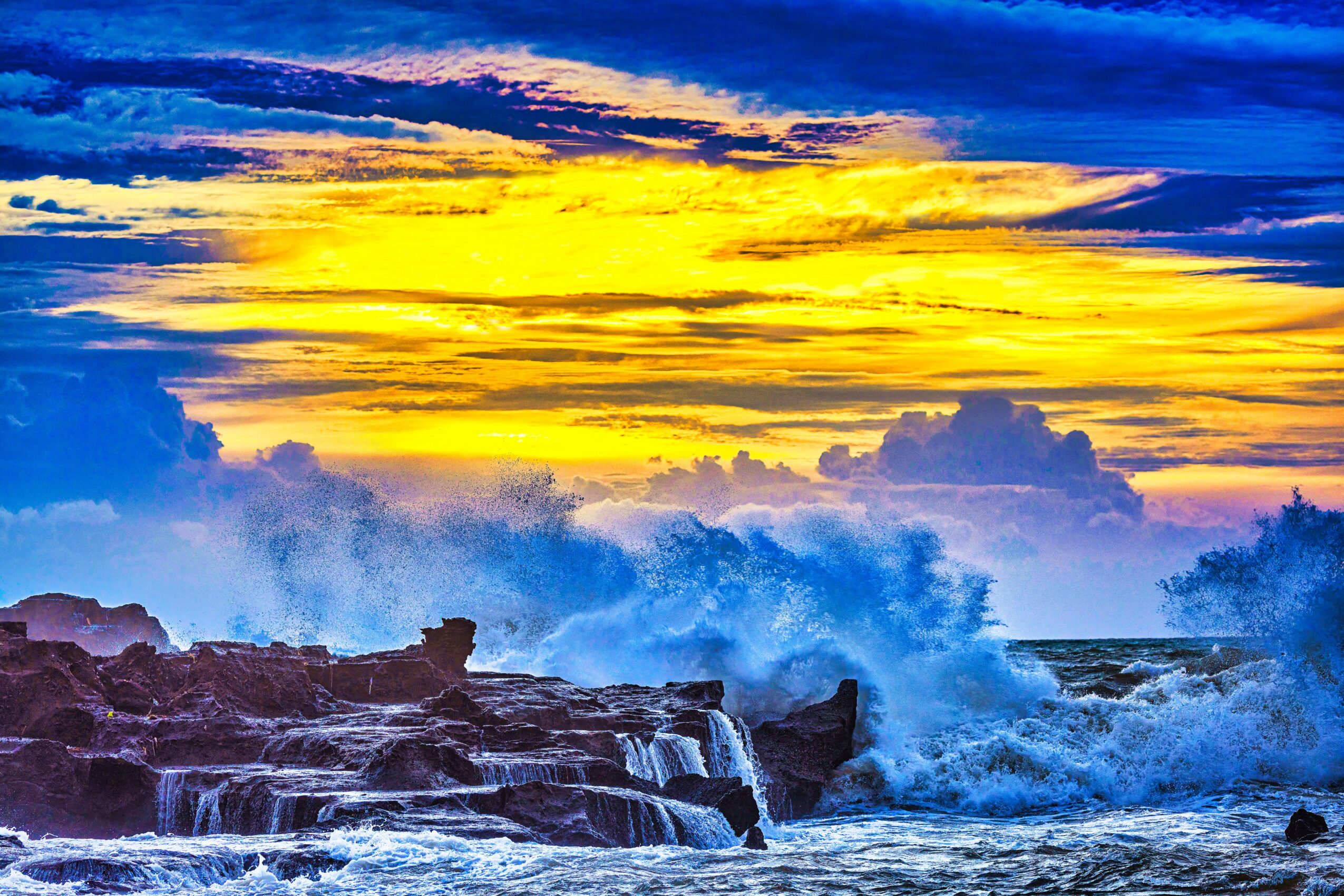 Vivid sunset sky with yellow, orange, and blue clouds over ocean waves crashing against rocks.