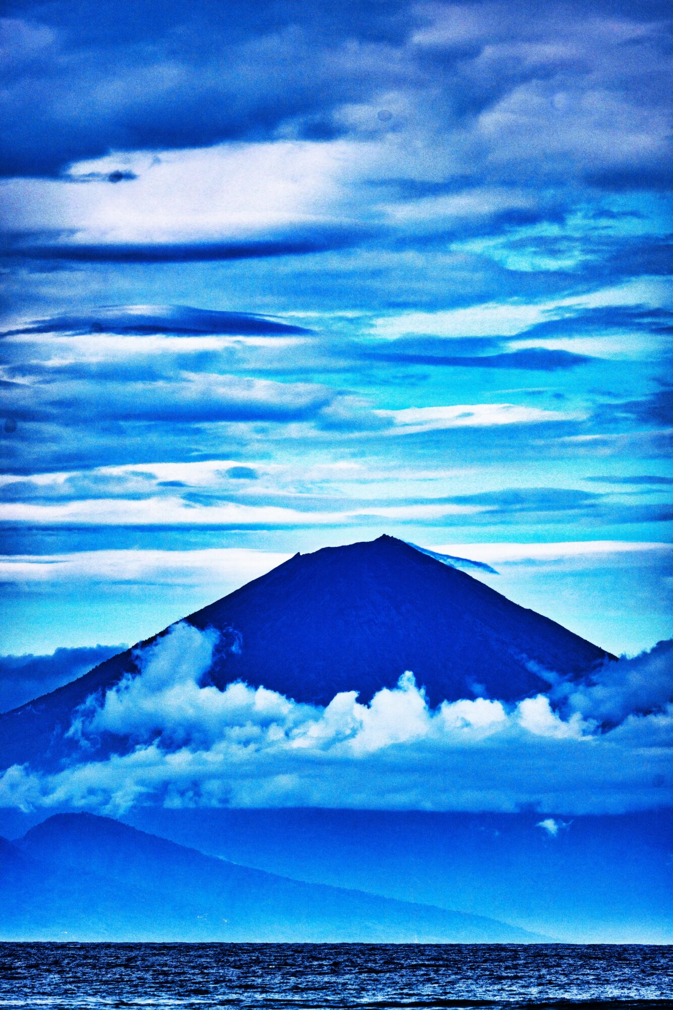 Mountain with clouds below and a sky with layered clouds above, in shades of blue and white.