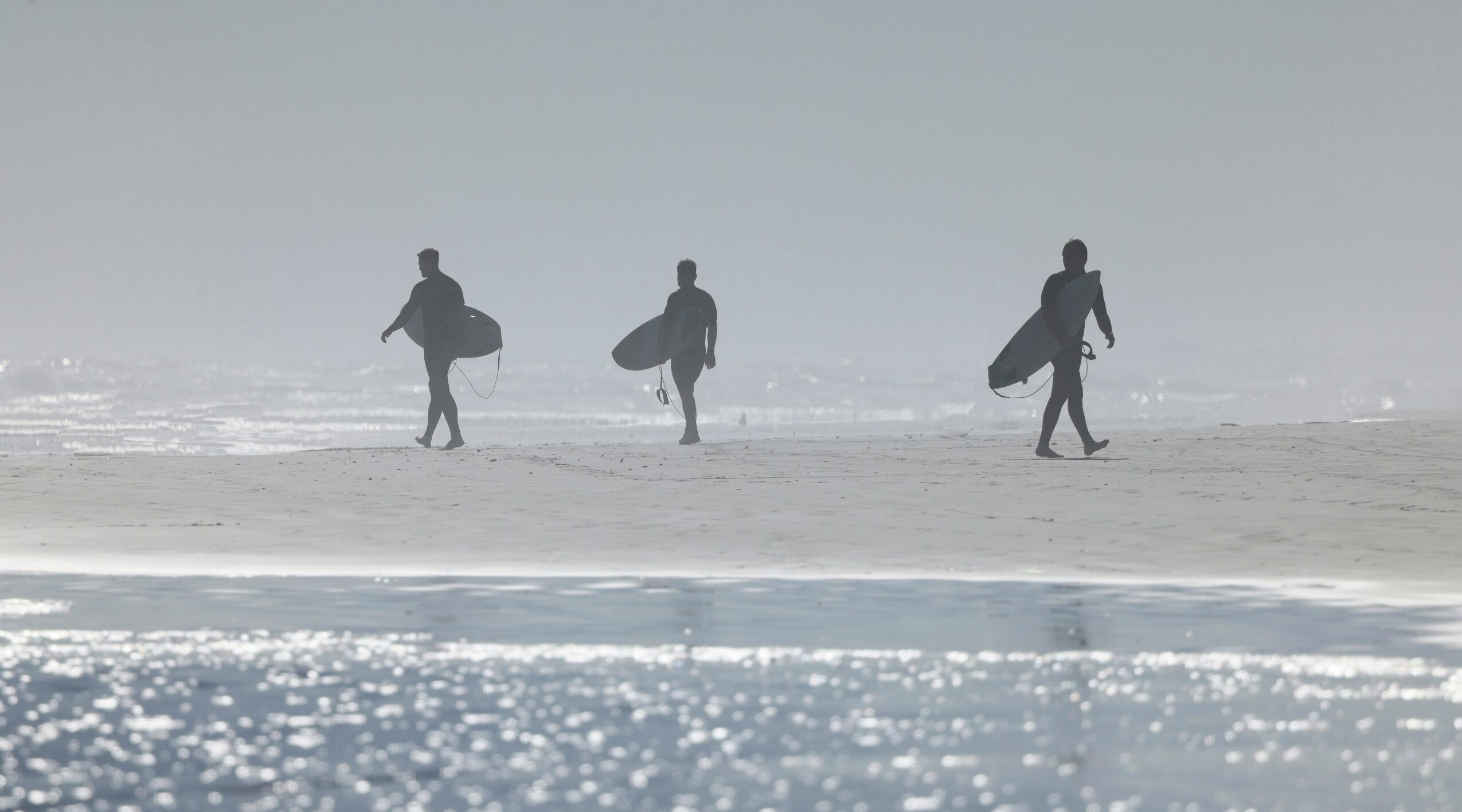 Three people walking on a beach carrying surfboards, with water and sand visible, in a foggy setting.