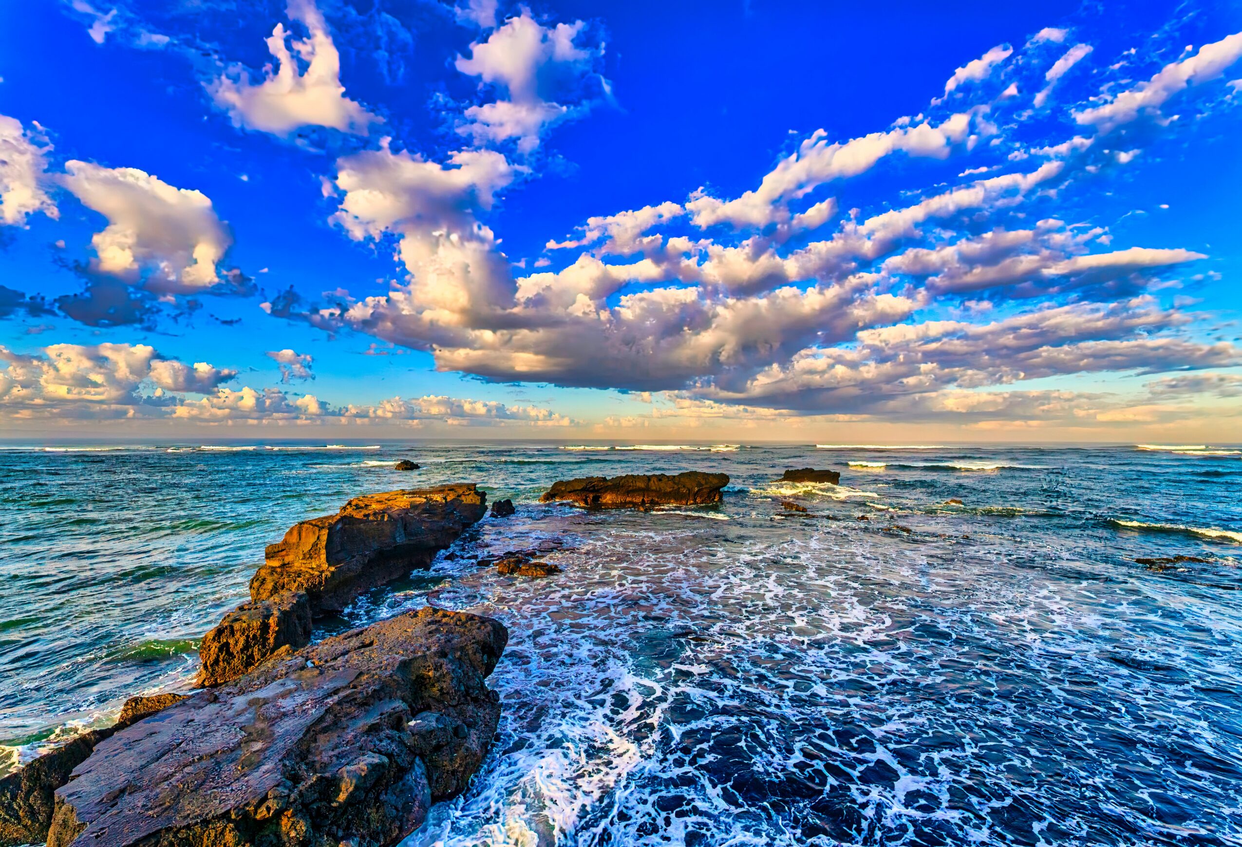 Ocean with rocks and a sky filled with clouds, blue water, and sunlight, under a partly cloudy sky.