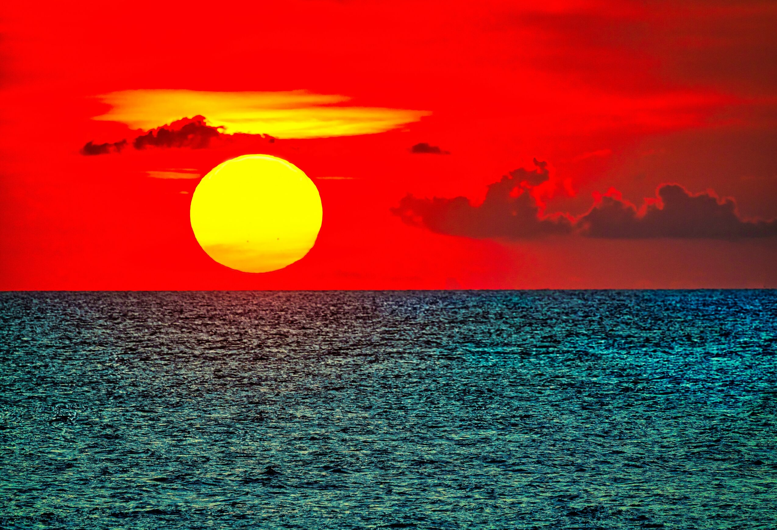 Sunset over the ocean with a large yellow sun and red sky, clouds in the background.