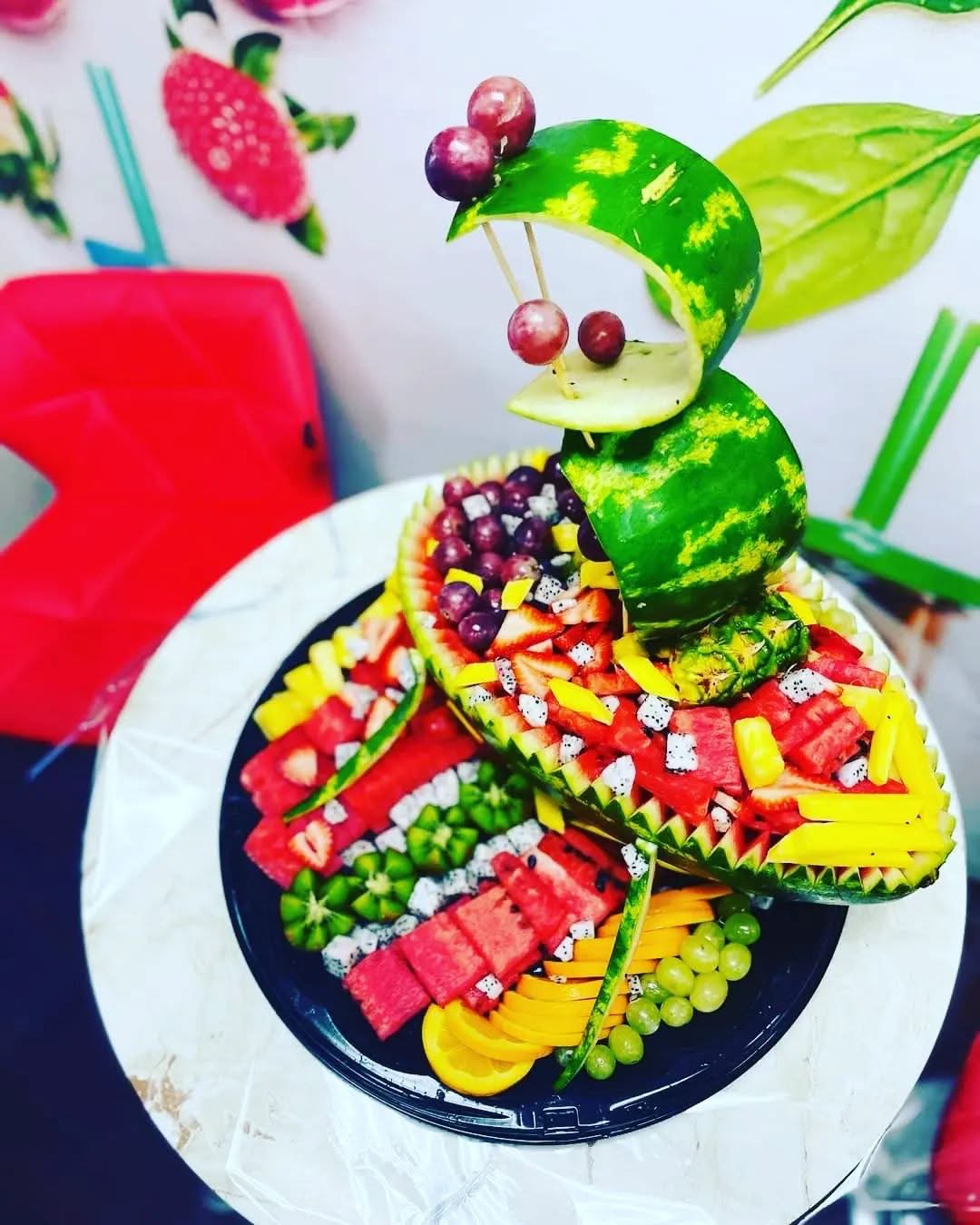 Fruit sculpture with watermelon, grapes, and other fruits arranged creatively on a black plate, with a white background and red chair.
