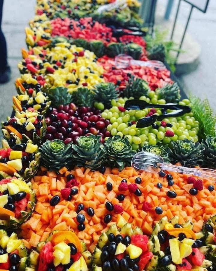 Display of various fresh fruits arranged in rows, including melons, grapes, and berries, at a market stall.