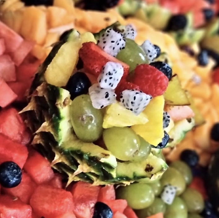 Pineapple half filled with mixed fruit including grapes, watermelon, dragon fruit, and pineapple chunks, surrounded by other fruit pieces.