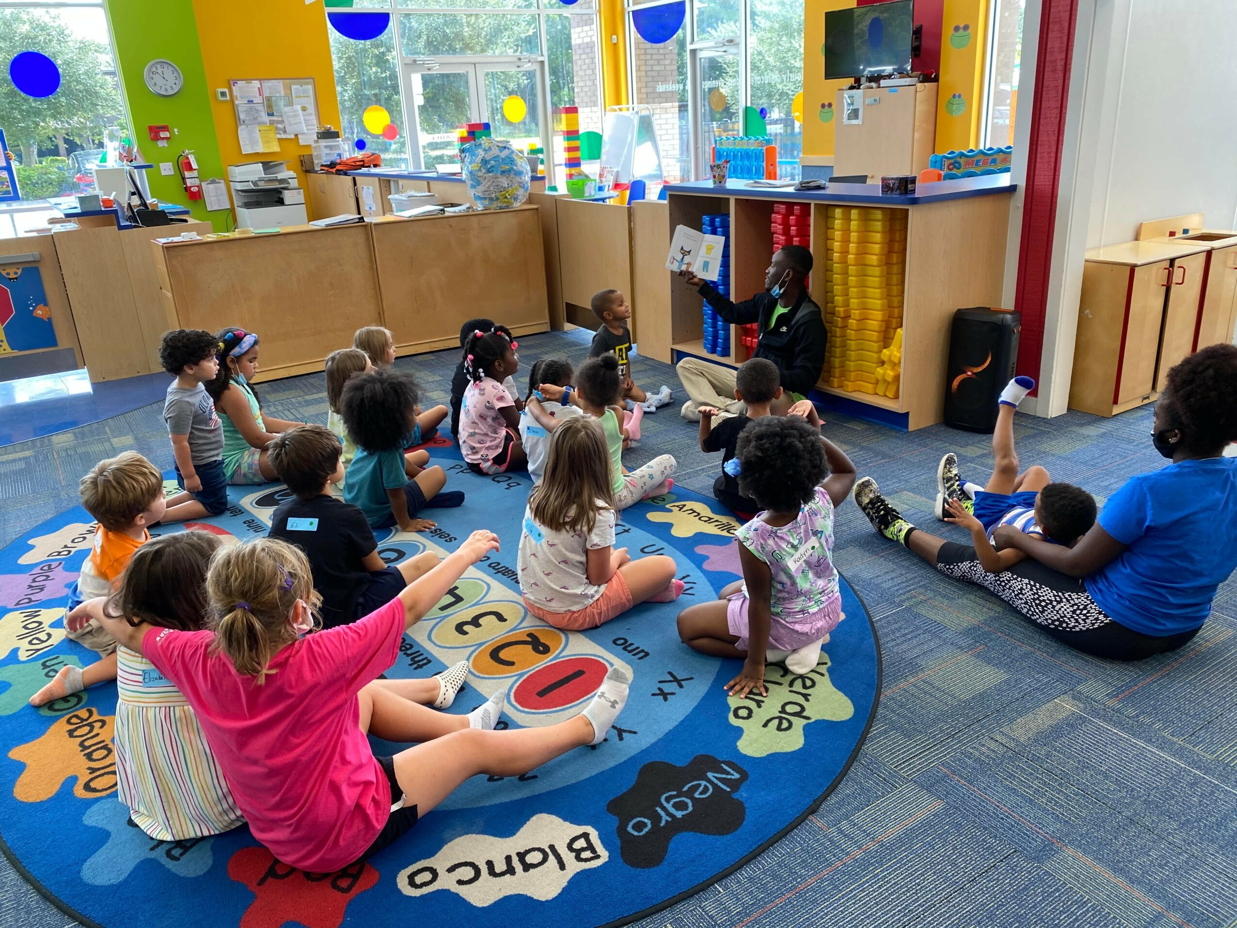 Children sitting on a colorful circular rug in a classroom, participating in a group activity with a teacher, bright windows in background.