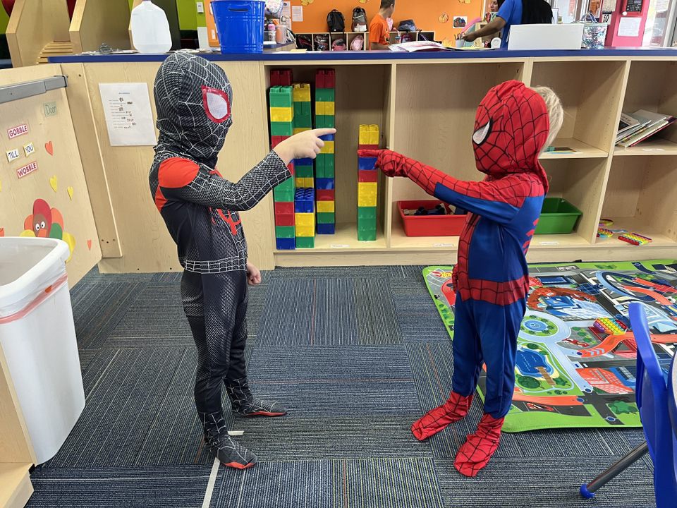 Two children dressed as Spider-Man and Miles Morales are playing together, touching hands in a colorful playroom.