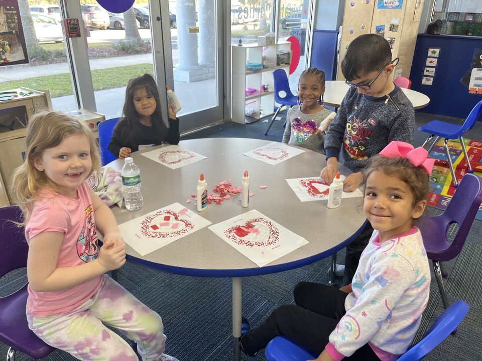 Five children sit around a table with art projects, smiling and engaging in a classroom activity.