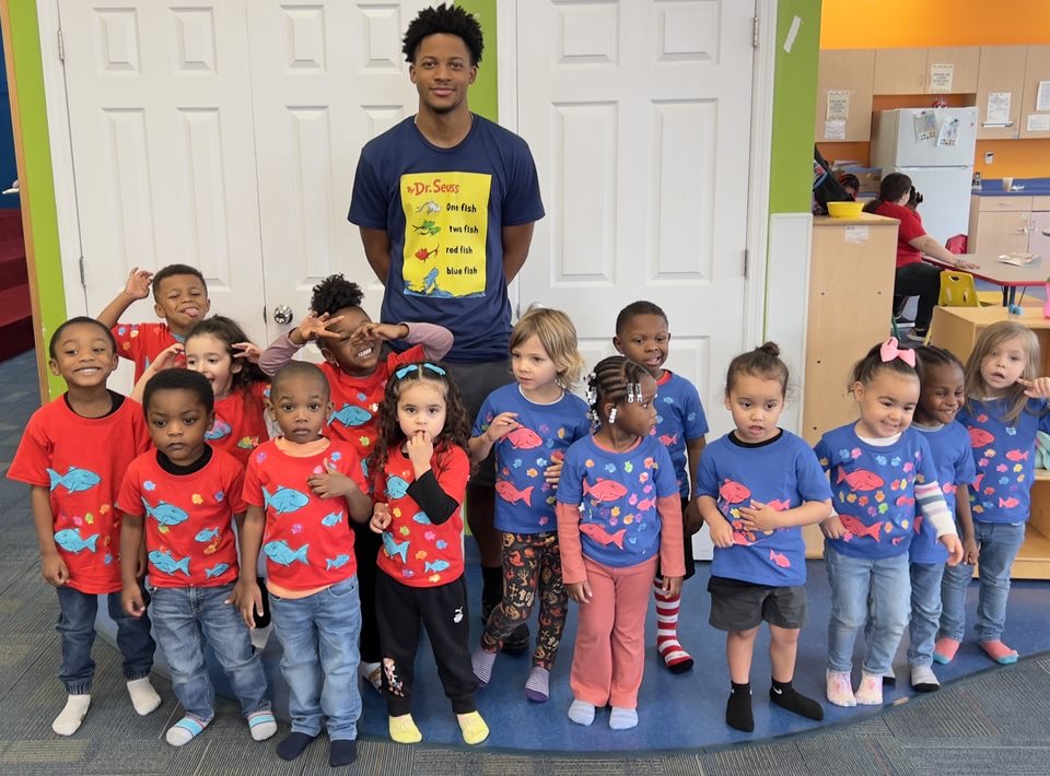 Group of young children and an adult in a classroom, children wearing matching blue and red shirts, standing in front of a white wall.