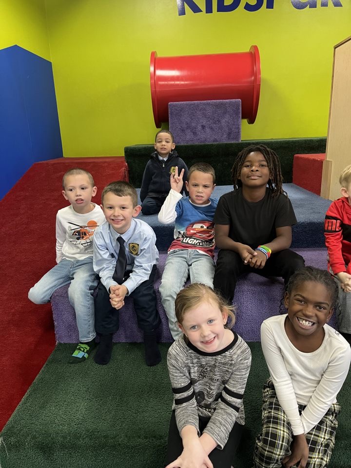 Group of children sitting on a purple platform in a colorful indoor play area, smiling and waving.