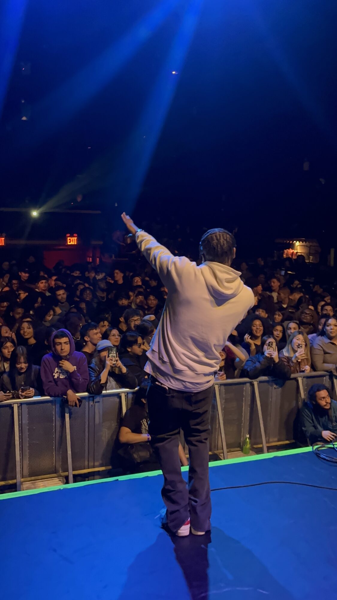 Performer on stage with back to audience, pointing upward, crowd watching attentively, dark background, blue stage floor.