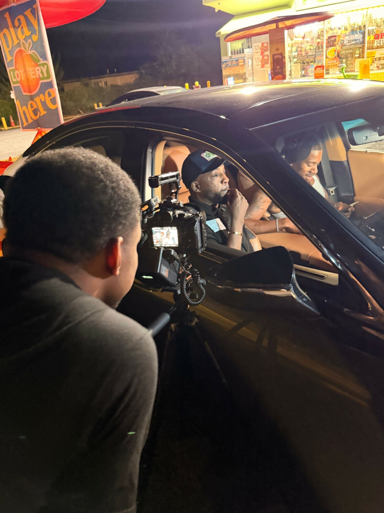Person with short hair leaning into a car at night, with a person inside the car, and a brightly lit store in the background.