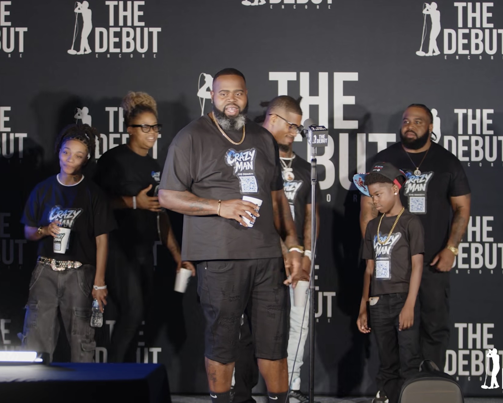 Group of people on stage at an event, some holding microphones and drinks, with a backdrop reading 'THE DEBUT'.