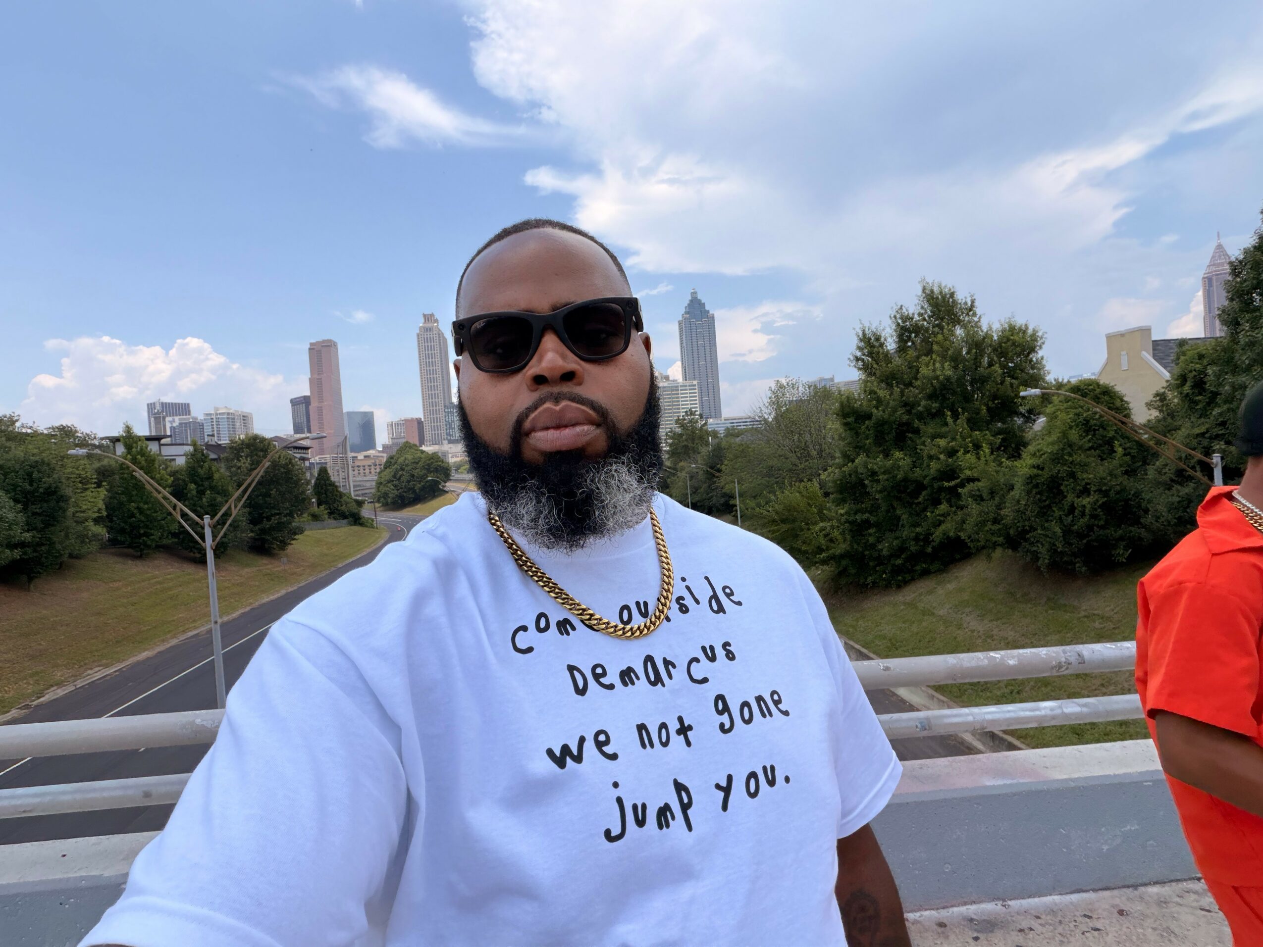 Man with sunglasses and beard wearing a white shirt with text, standing outdoors with city skyline and trees behind him.