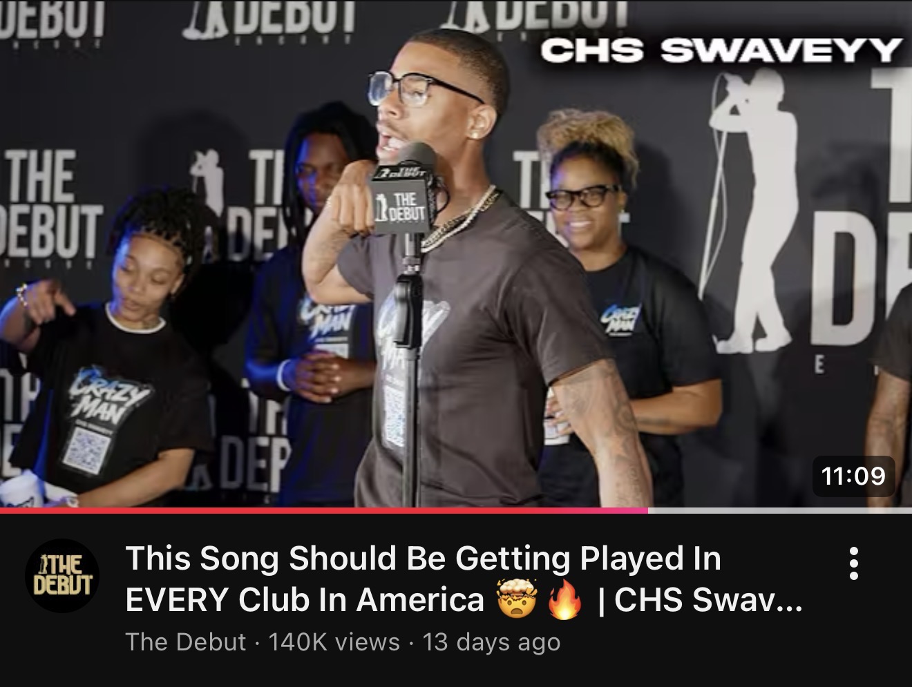 Young man with glasses speaking into microphone on stage with four people behind him, black background, event banner, 11:09 timestamp.