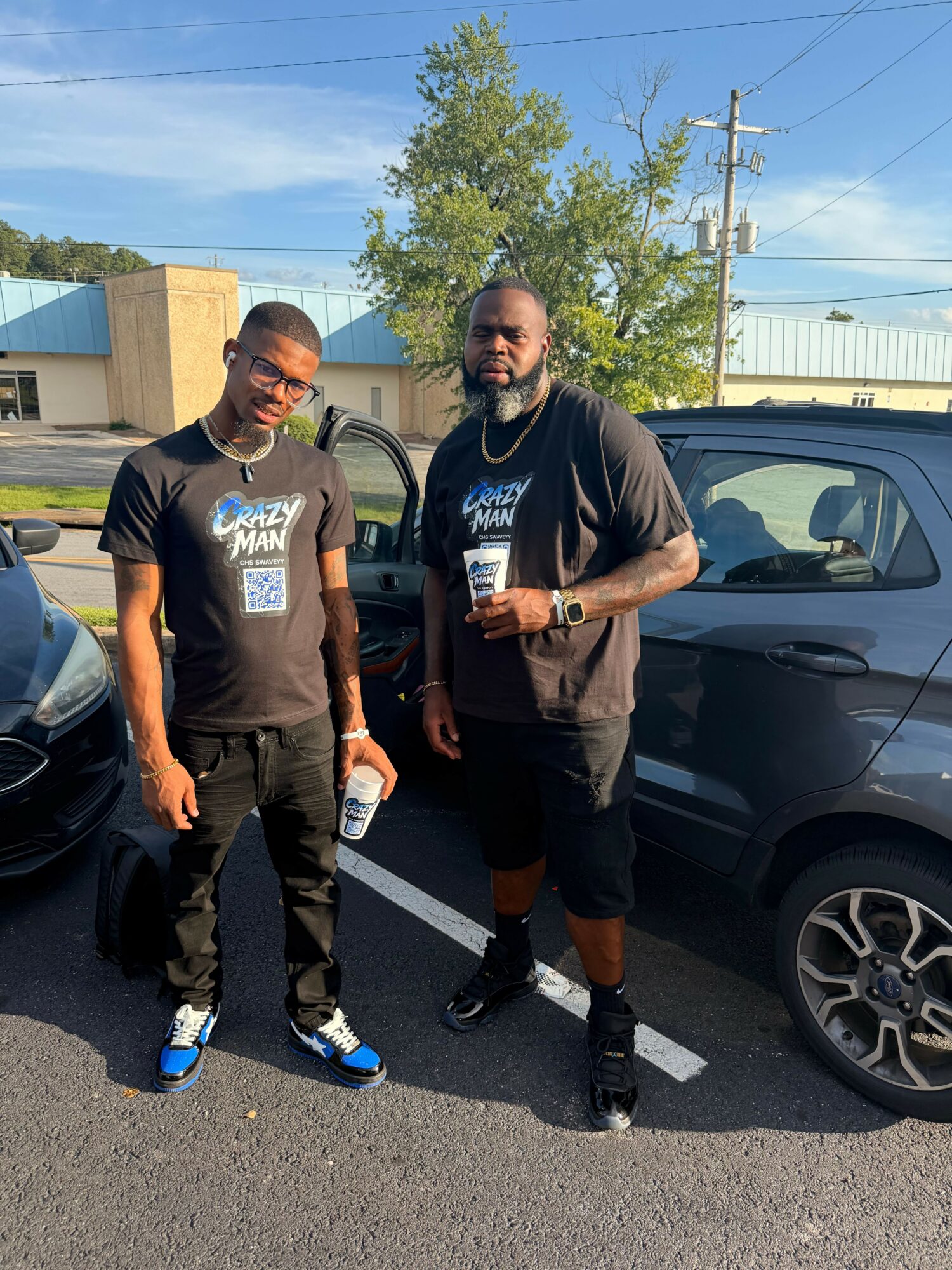 Two men standing next to a parked car in a parking lot, with trees and a building in the background.