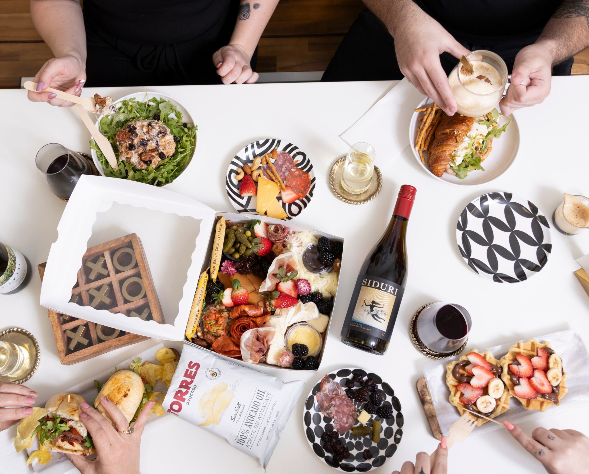 People gathered around a table with desserts, wine, and snacks, viewed from above. The table has various plates and glasses.