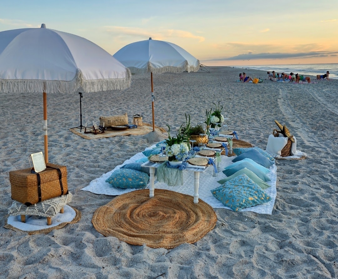 Beach setup with a low table, cushions, umbrellas, and decorative plants on sand, with people and ocean in background.