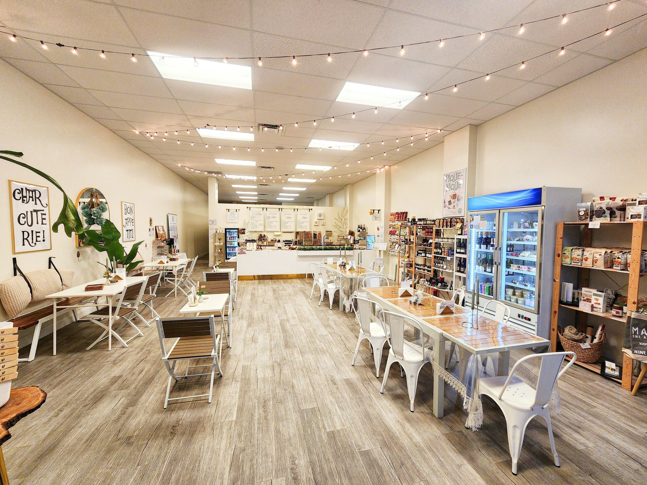 Empty store with tables, chairs, shelves, and a refrigerator, illuminated by ceiling lights, with a wooden floor and white walls.