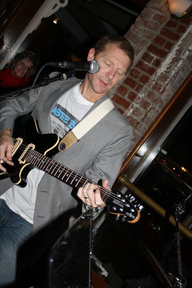 Man playing guitar and singing into microphone in a brick-walled venue, with a woman in the background.