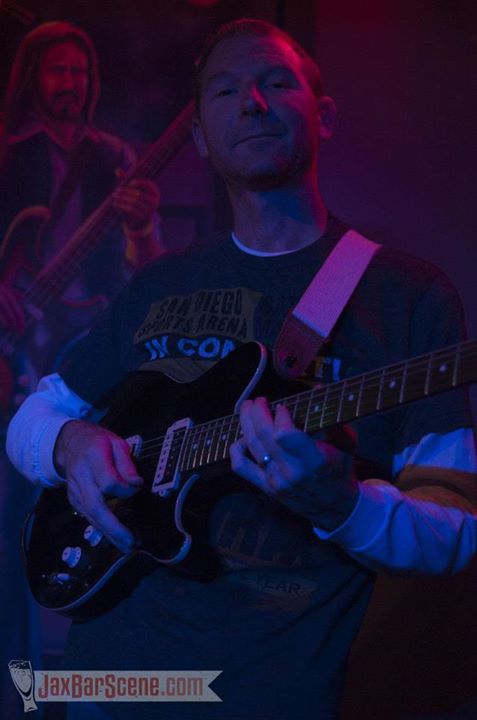 Person playing an electric guitar on stage with another musician in the background, under dim lighting.