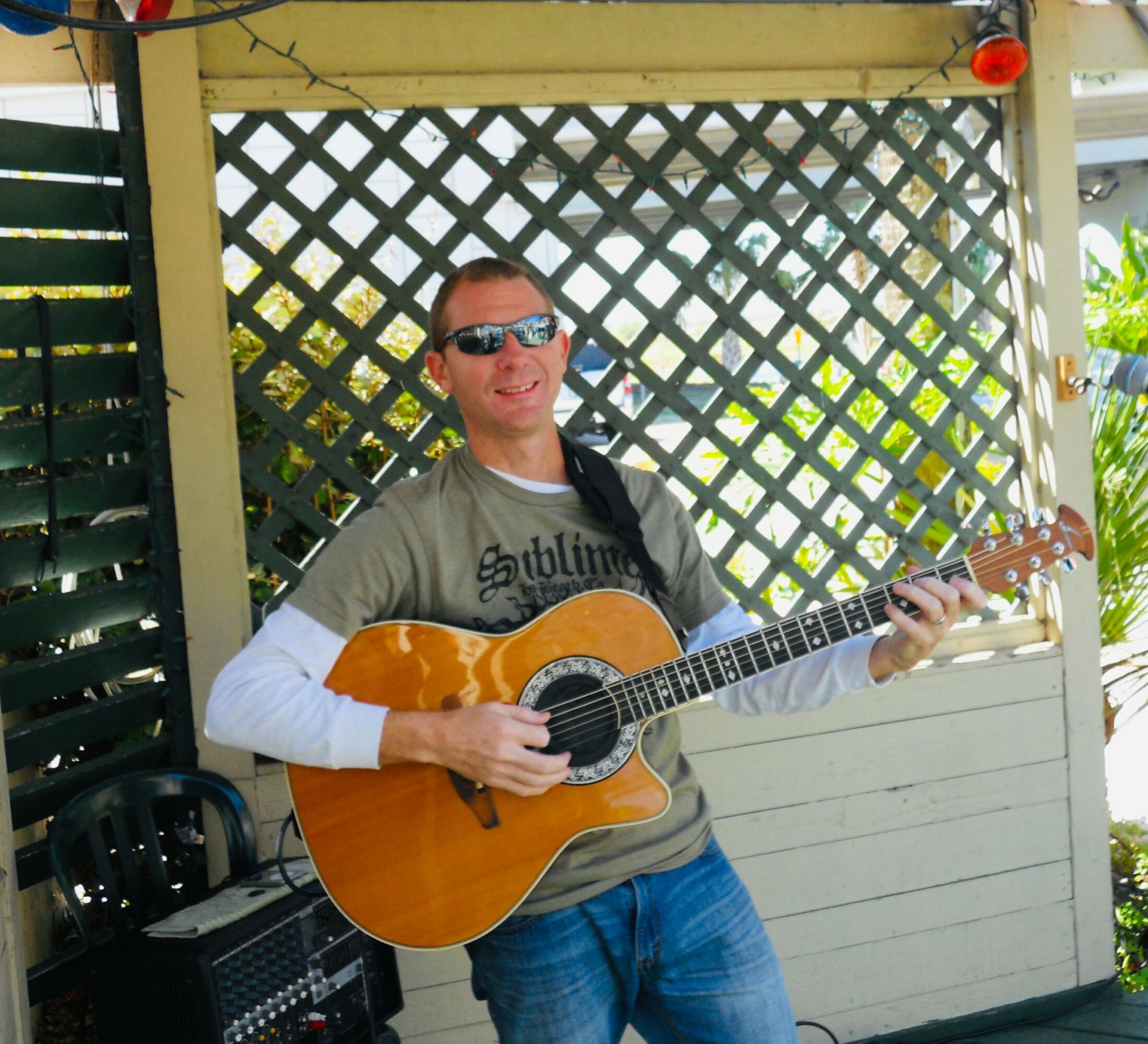 Person wearing sunglasses playing an acoustic guitar outdoors, standing in front of a lattice fence.