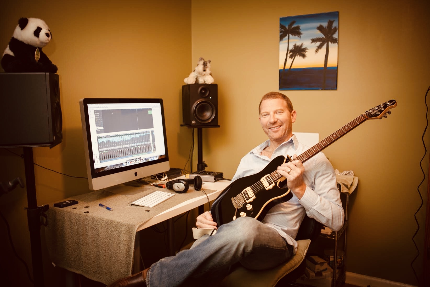 Man sitting in a room holding an electric guitar, smiling, with music editing software on a computer screen behind him.