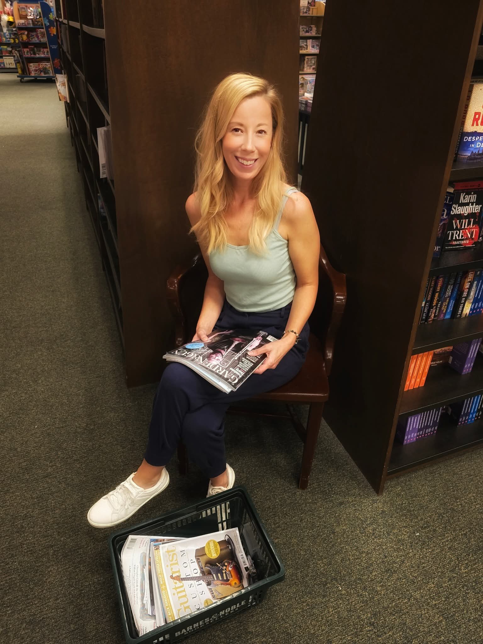 Woman with blonde hair sitting on a chair in a bookstore, holding a magazine, smiling, with bookshelves nearby.