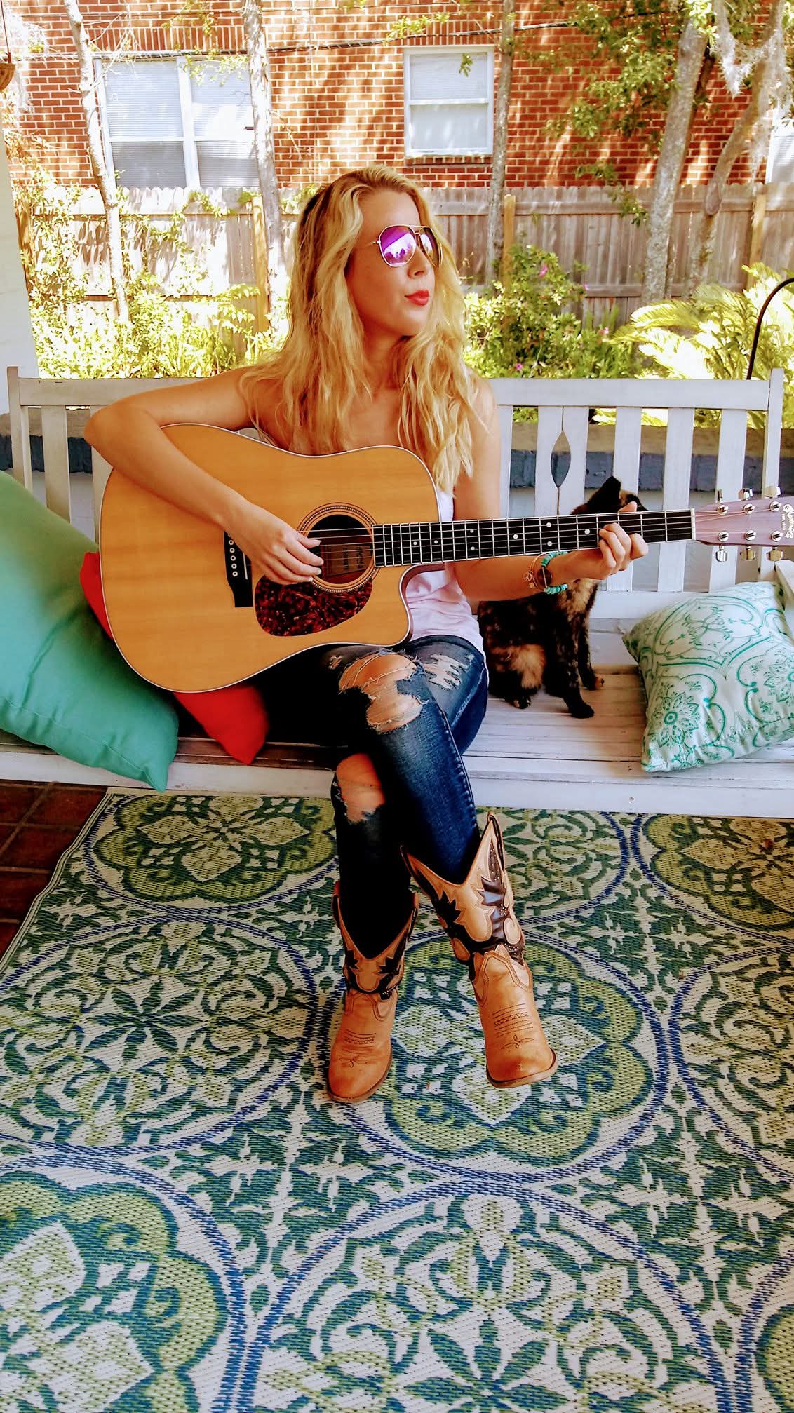 Woman with long hair and sunglasses playing an acoustic guitar on a porch, wearing ripped jeans and boots.