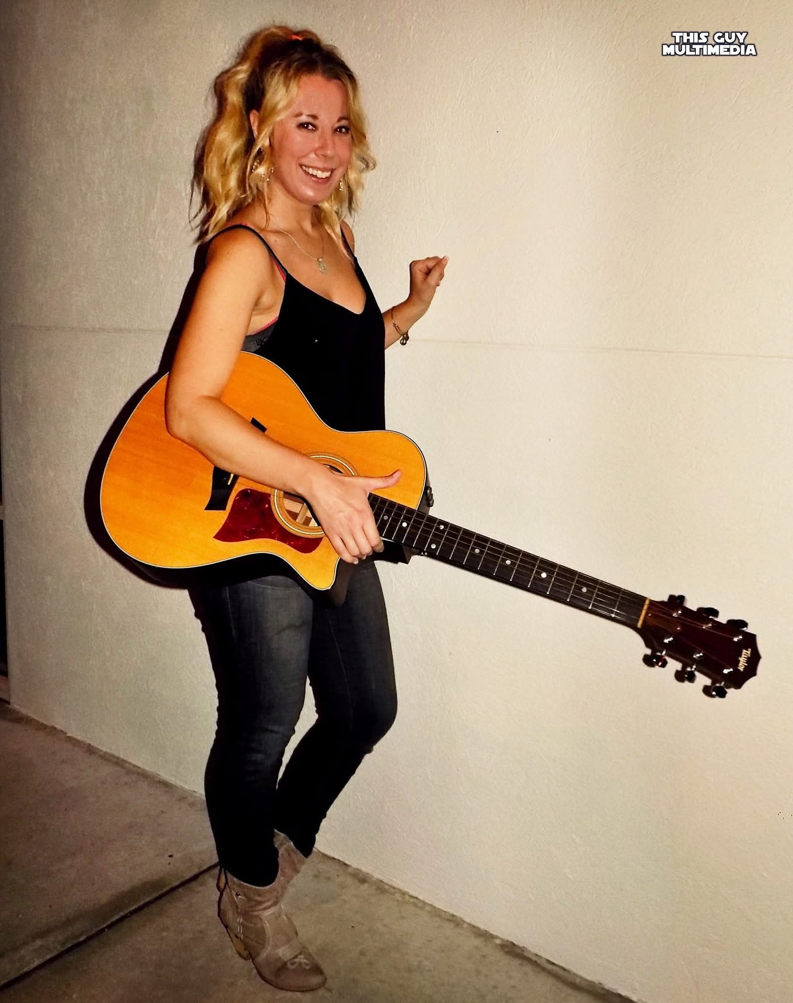 Woman with wavy blonde hair holding an acoustic guitar, smiling, standing against a plain wall, wearing a black top and jeans.