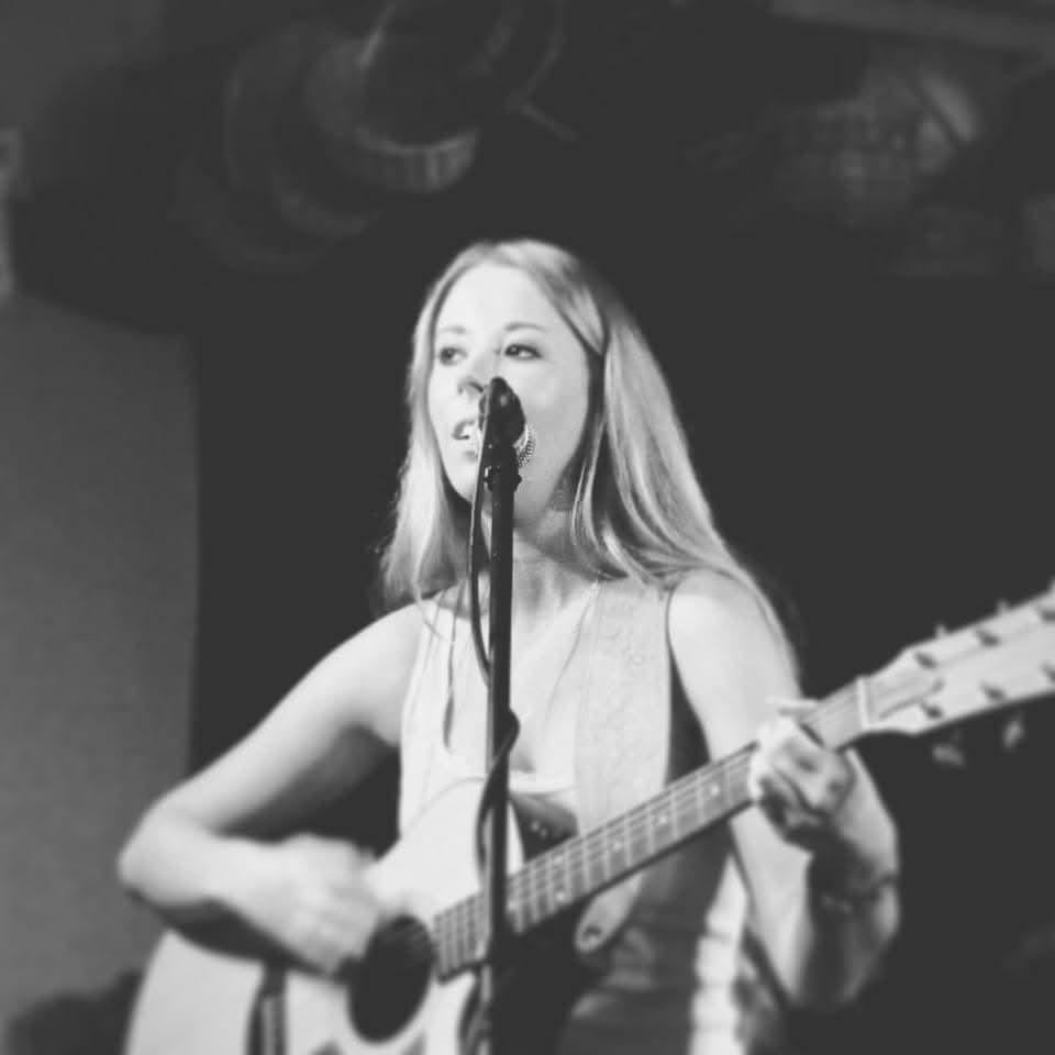 A woman with long hair playing an acoustic guitar and singing into a microphone on stage.