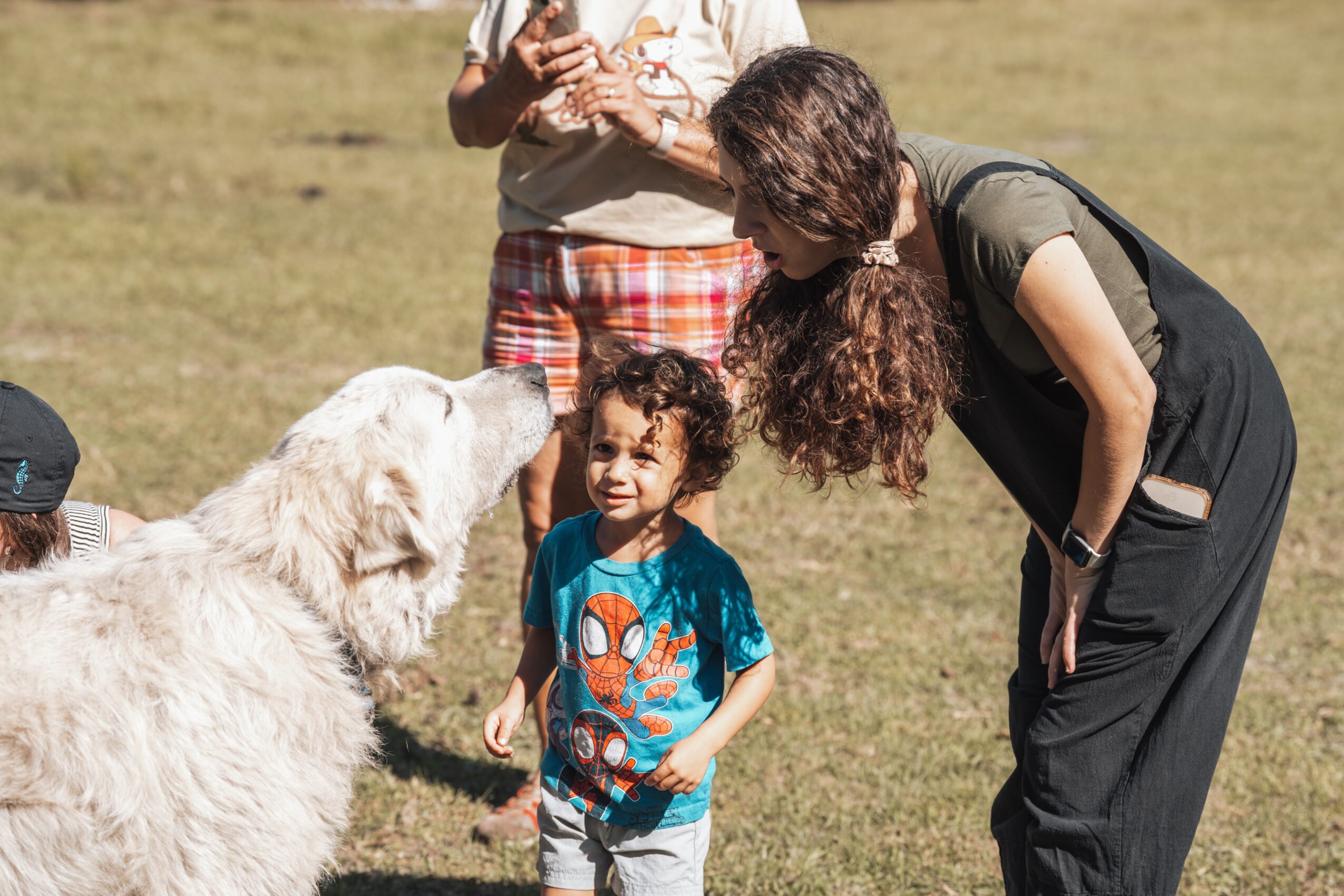 Woman bending down to talk to a young girl with curly hair, outdoors on grass, with a large white dog nearby.
