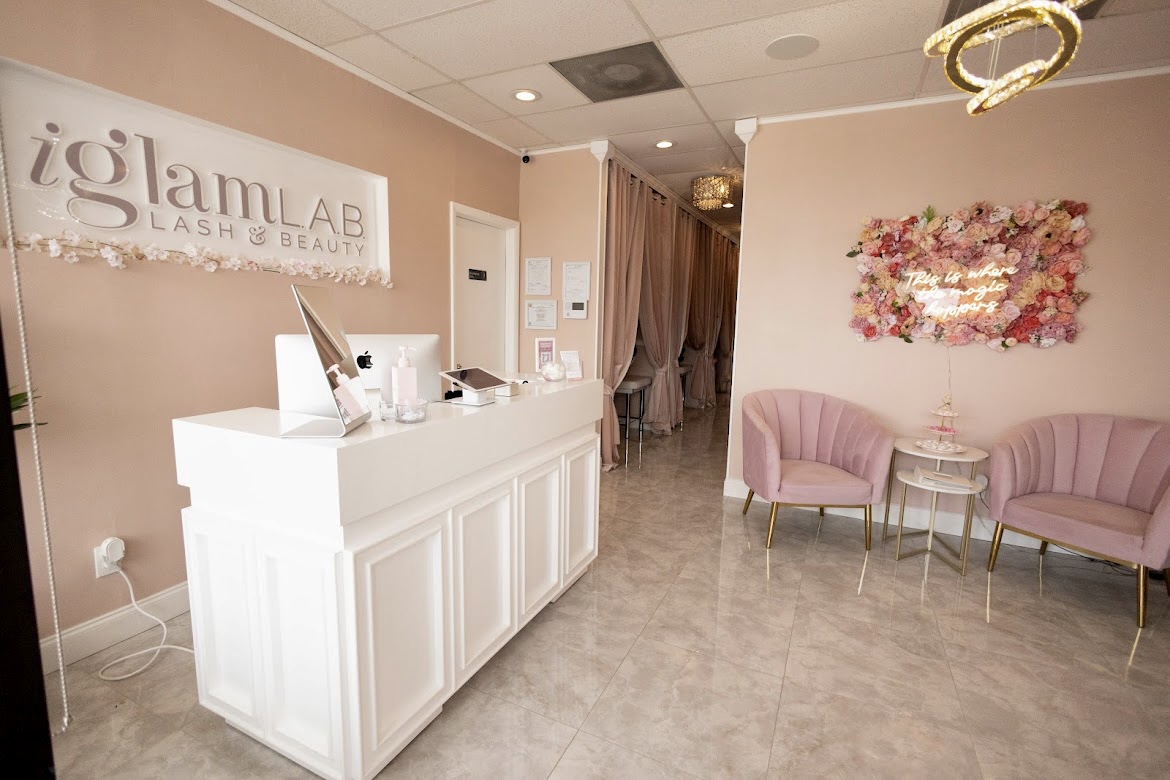 Reception area with a white counter, pink chairs, floral wall art, and a sign reading 'iglam LAB' on beige walls.