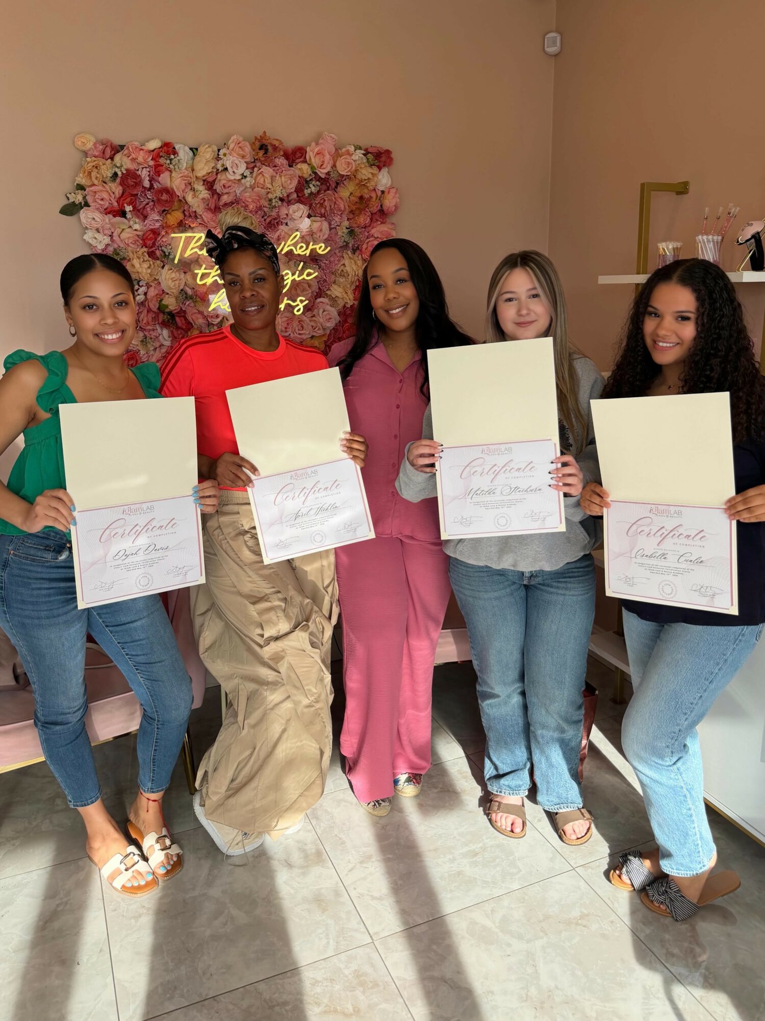 Five women standing indoors, holding certificates, smiling, with a floral backdrop behind them.