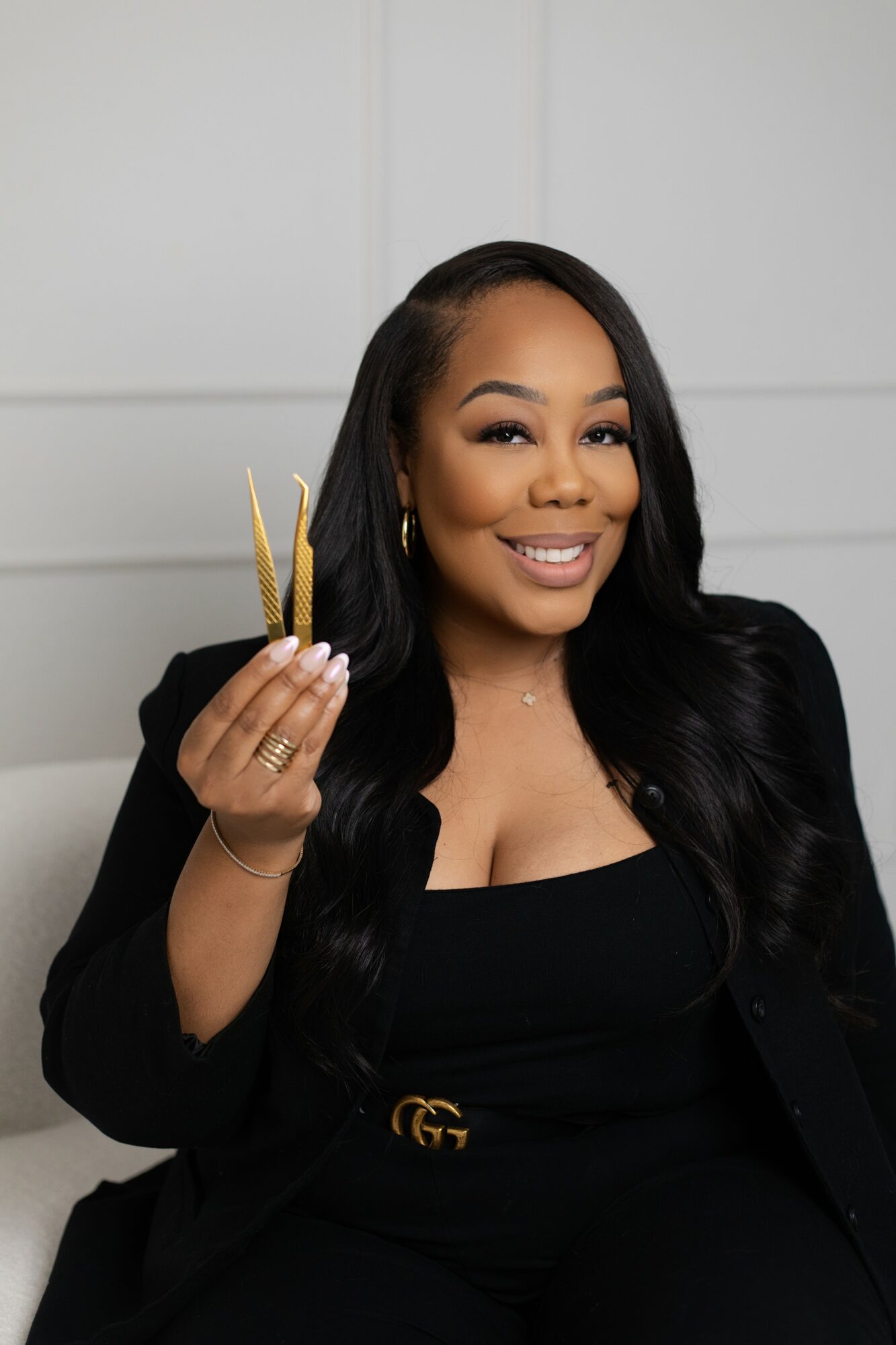 Woman with long black hair smiling, holding a small plate with food, dressed in black, in a neutral background.