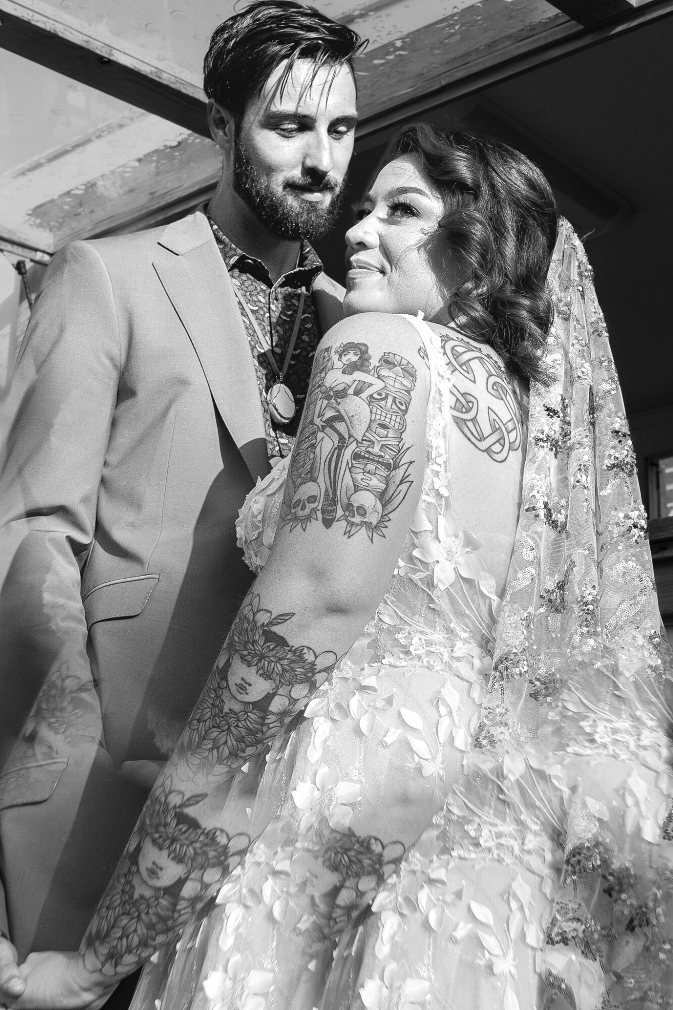 A couple dressed in wedding attire, the woman with tattoos on her arm, smiling and looking at each other.