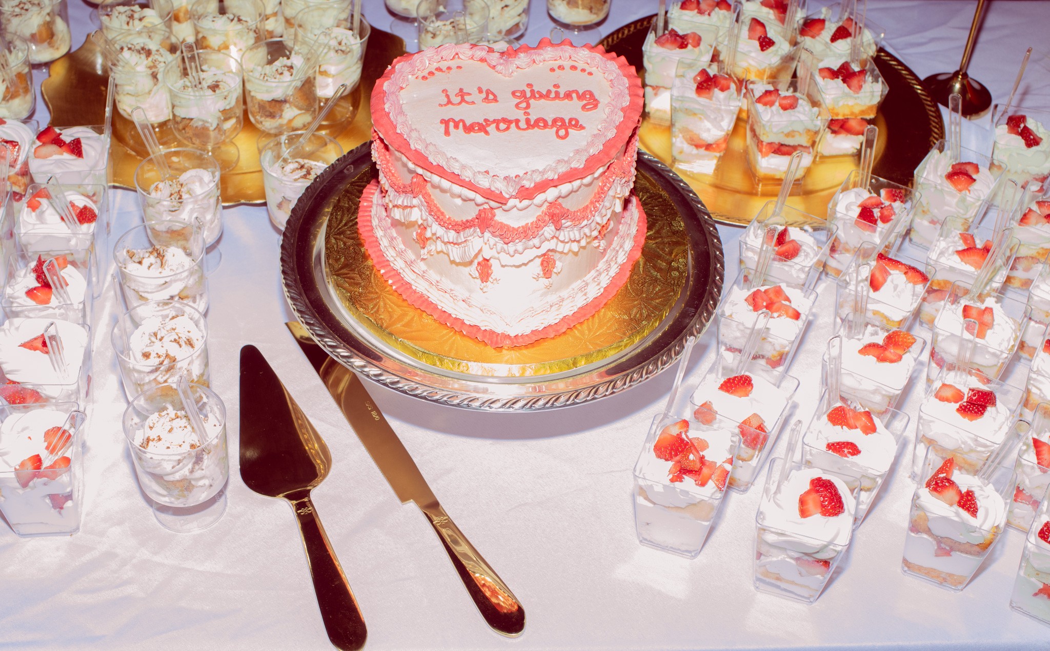 Heart-shaped cake with pink and white frosting, surrounded by small desserts with strawberries, on a table with utensils.