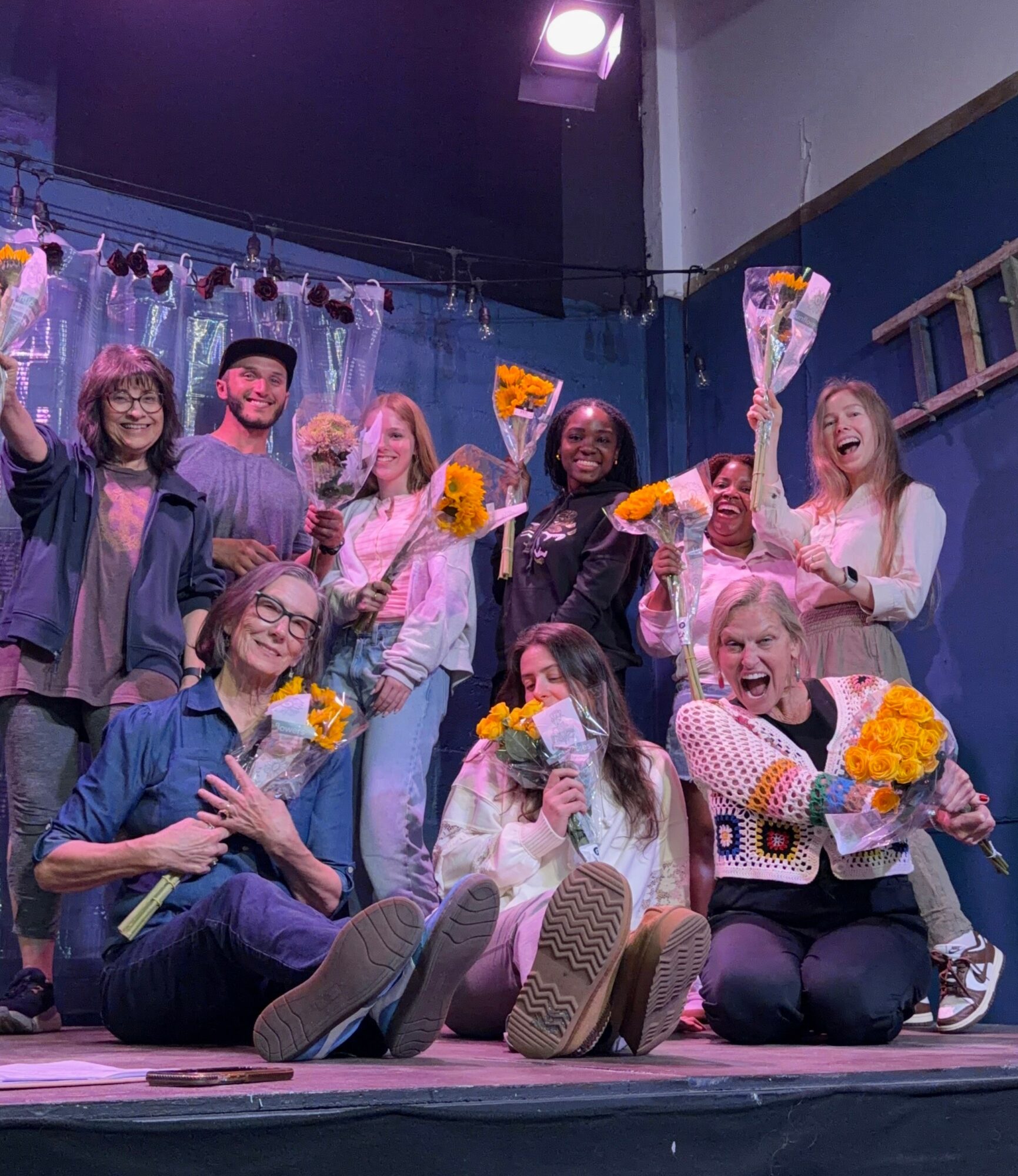 Group of nine people on stage holding flowers, smiling, and celebrating, with a curtain and stage equipment in background.