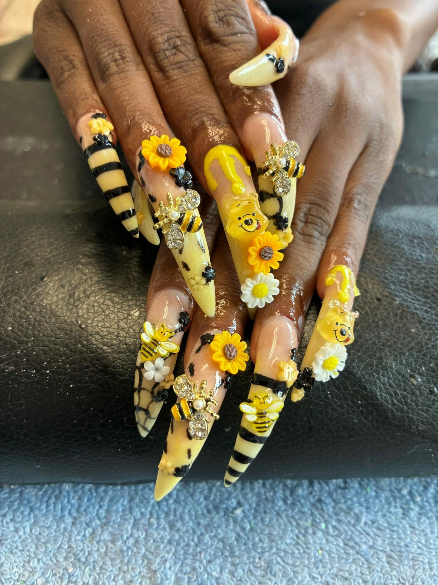 Hands with decorated long nails featuring yellow, black, and white striped patterns, adorned with small yellow and white flowers and bee accessories.