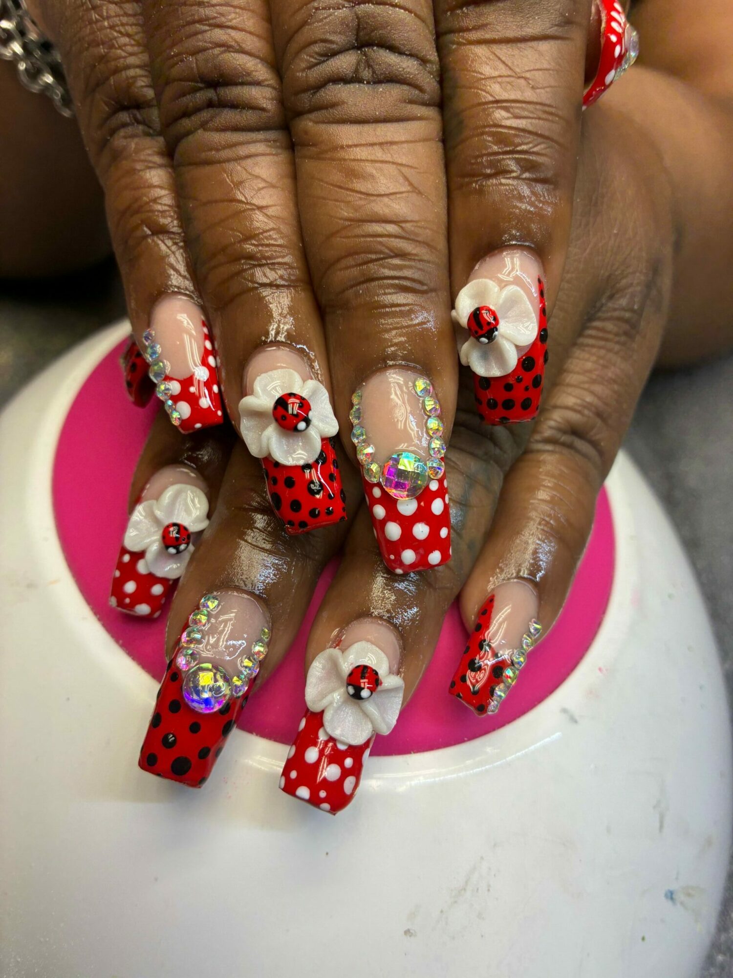 Close-up of hands with decorated nails featuring red, white, and black designs, including flowers and polka dots, on a pink and white device.