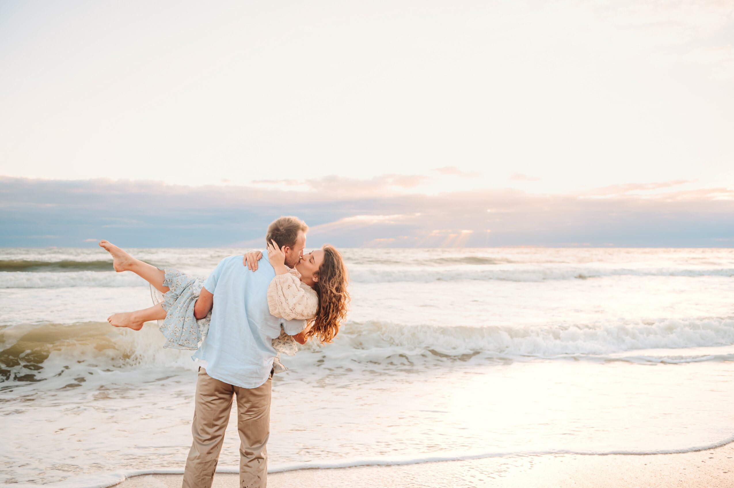 Person holding a child on the beach with waves and sky in the background, sunset lighting, calm scene