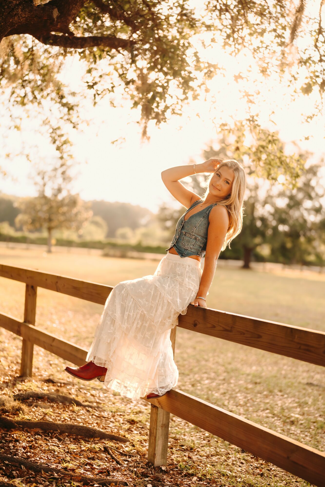Woman sitting on a wooden fence outdoors during sunset, wearing a long white skirt and a sleeveless top, with trees in the background.