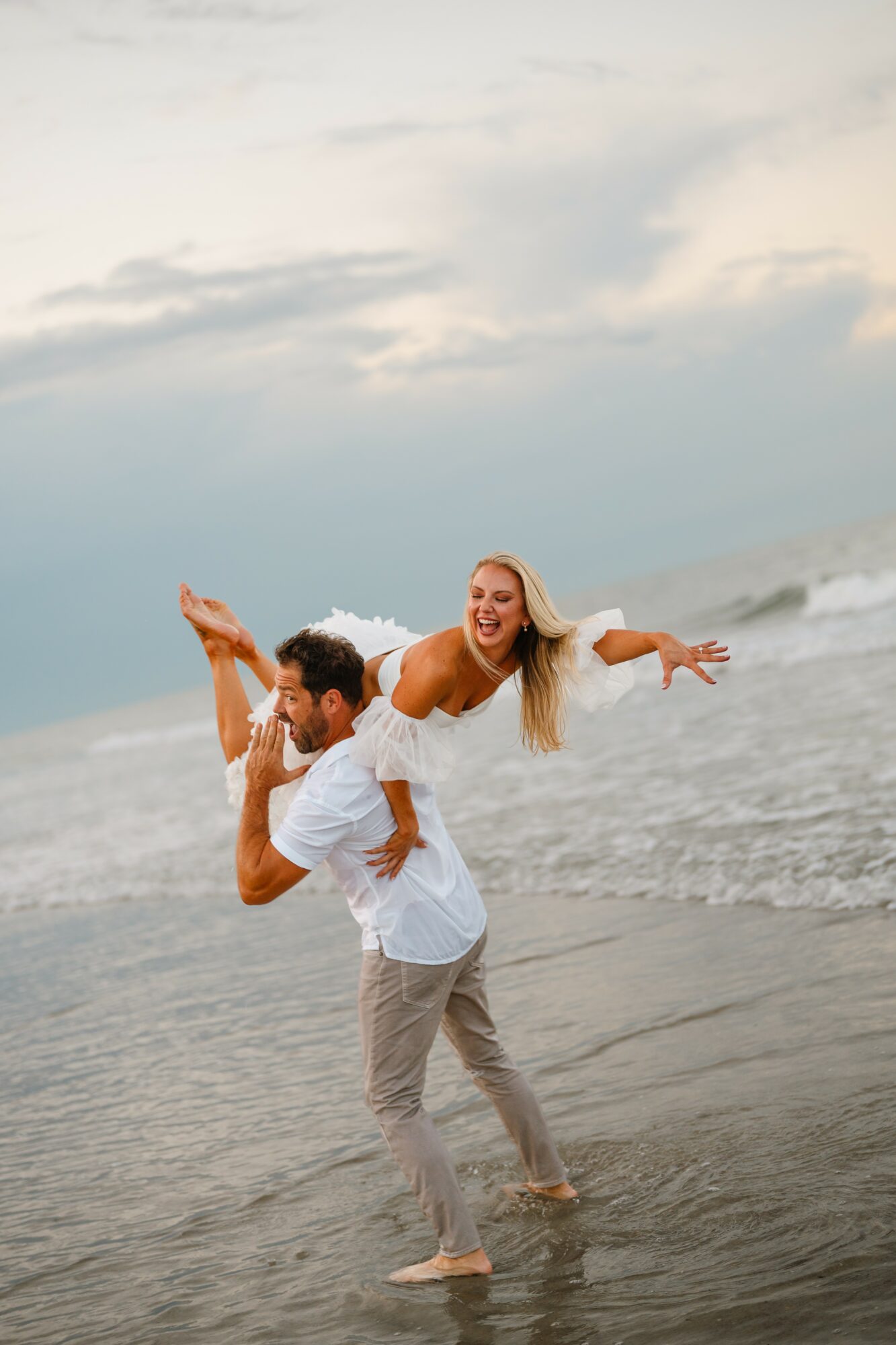 A man carries a woman on his shoulders at the beach, both smiling and enjoying the water and sky.