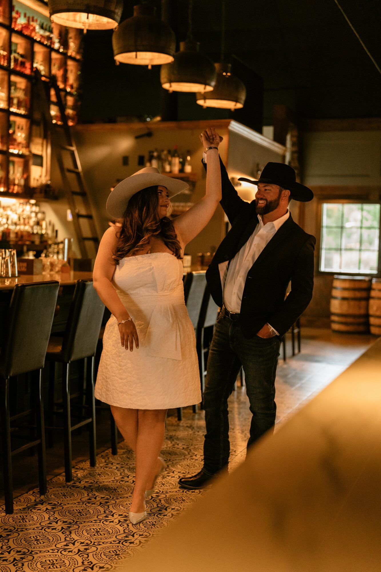 Two people dance in a dimly lit bar, holding hands, with shelves of bottles behind them.
