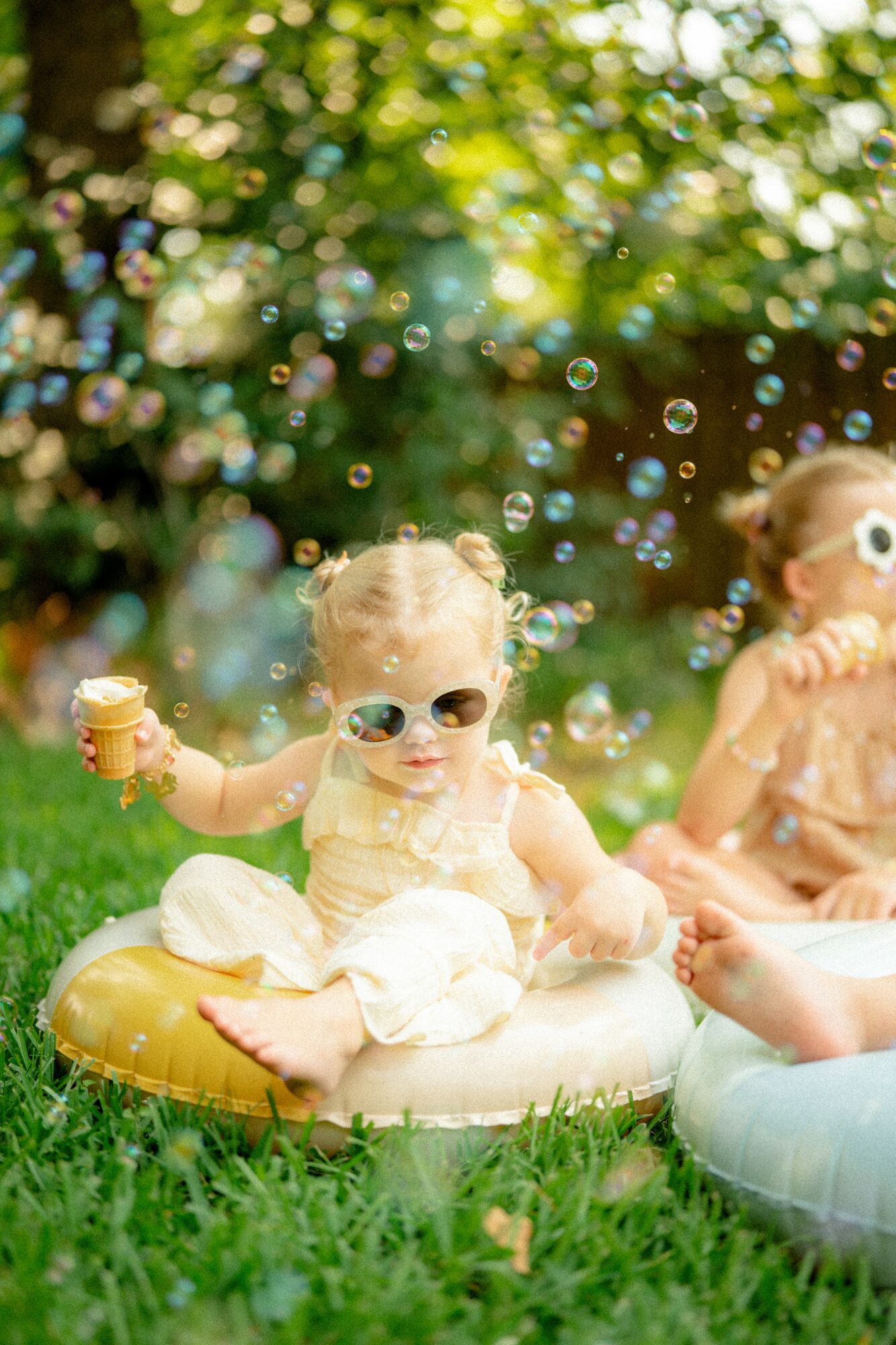Two children sitting on inflatable rings outdoors, one holding an ice cream, surrounded by colorful bubbles and greenery.