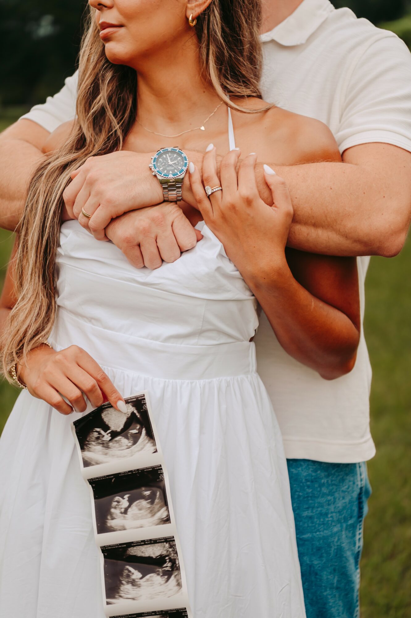 Person in white dress holding ultrasound images, with another person embracing from behind, outdoors.
