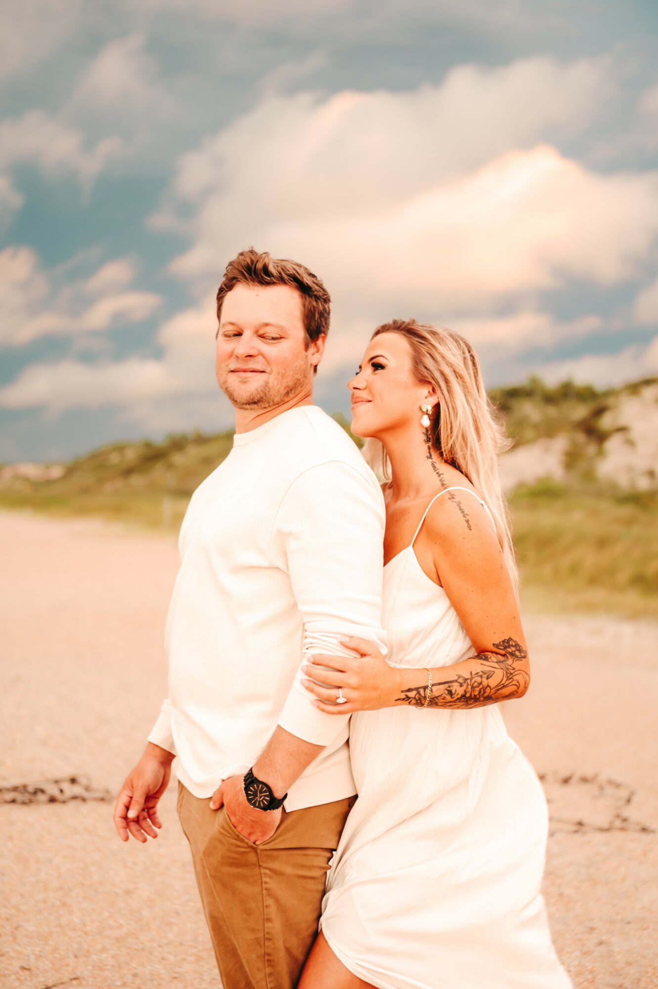 A couple standing outdoors on a beach with a cloudy sky, the woman hugging the man from behind, both smiling.