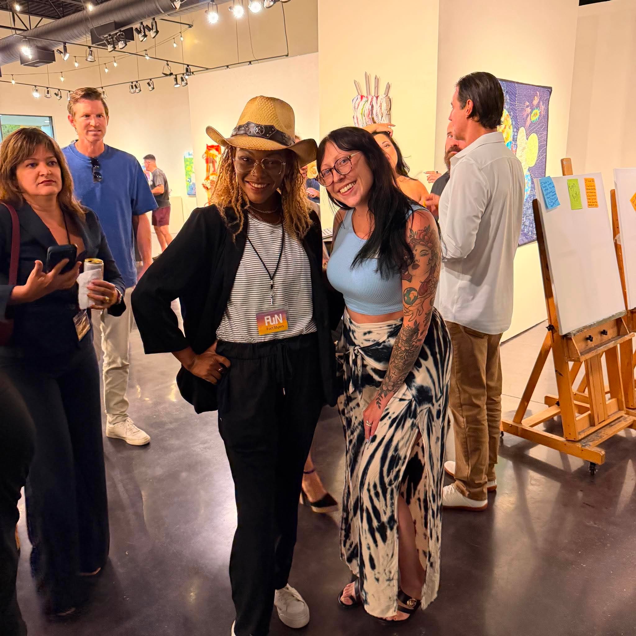 Two women smiling and posing together at an art gallery event, surrounded by other people and artwork.