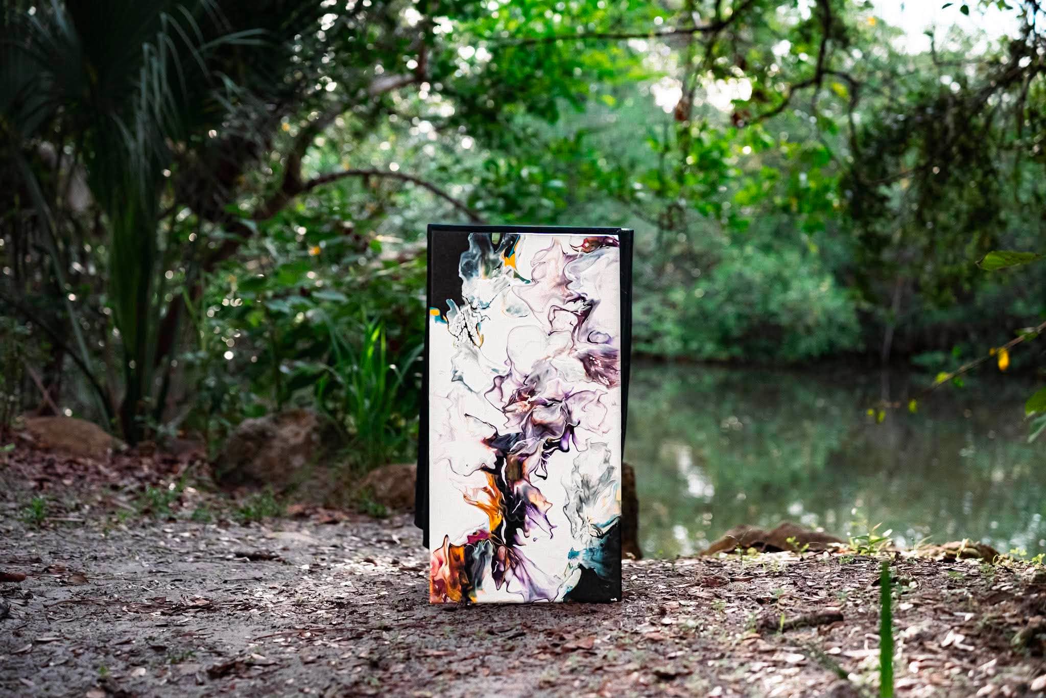 A tall, abstract painting on a canvas stands on the ground near a pond surrounded by trees and greenery.